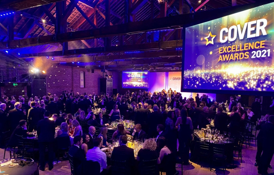 An awards ceremony photographed, Cover Excellence Awards 2021. People are stood at tables and sat down. The lighting is atmospheric, purple and yellow.