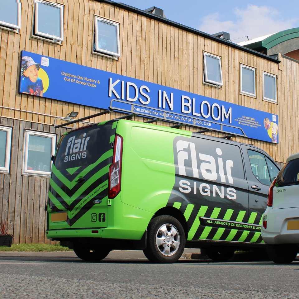 Flair Signs, a green and black van, outside a shop they have created the signage for. 'Kids in Bloom' on a blue bold background