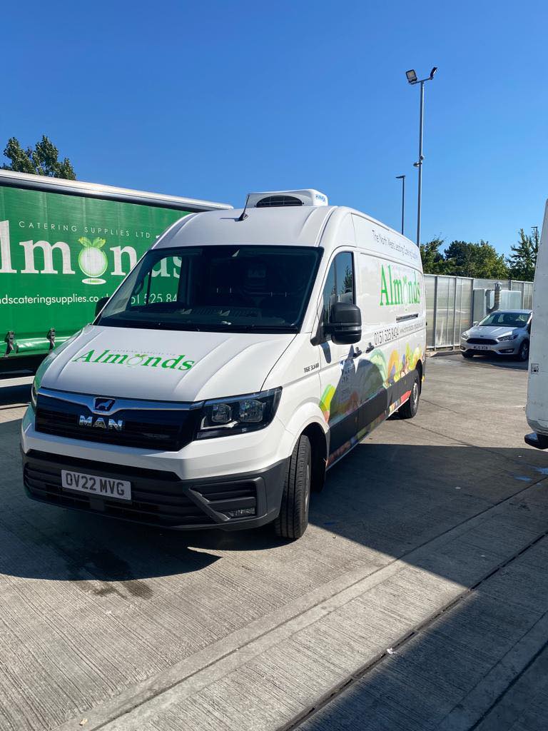 An image of an Almonds Catering Van. The van is white with their green logo on the front bonnet. The sky is blue and there is another larger van with the logo on behind the main van in focus.