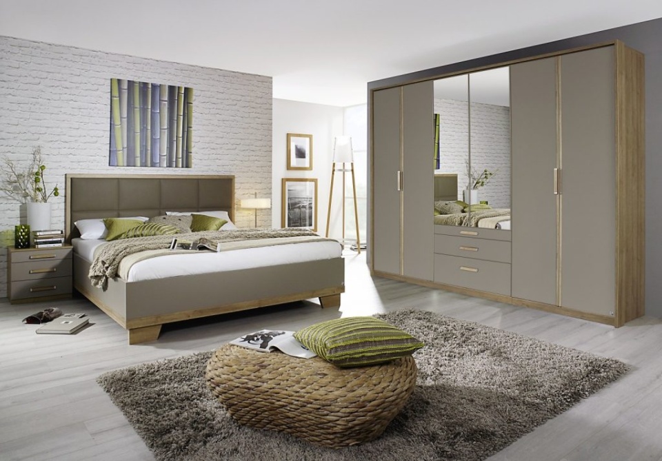 A bedroom is shown with a double bed on the left hand side. Everything is very stylised, with a green and brown colour scheme. The walls are neutral white colours, the light coming in at the back lights up the room with a glow. There is a wardrobe with a mirror in the centre, it is a large wardrobe. The bed itself has green and grey cushions on it, and a padded headboard. The image above the headboard is a canvas of bamboo. In front of the bed and at the bottom of the image there is a round wooden seat or stool with a green pillow on it. Underneath this there is a fluffy grey rug, and a wooden floor.